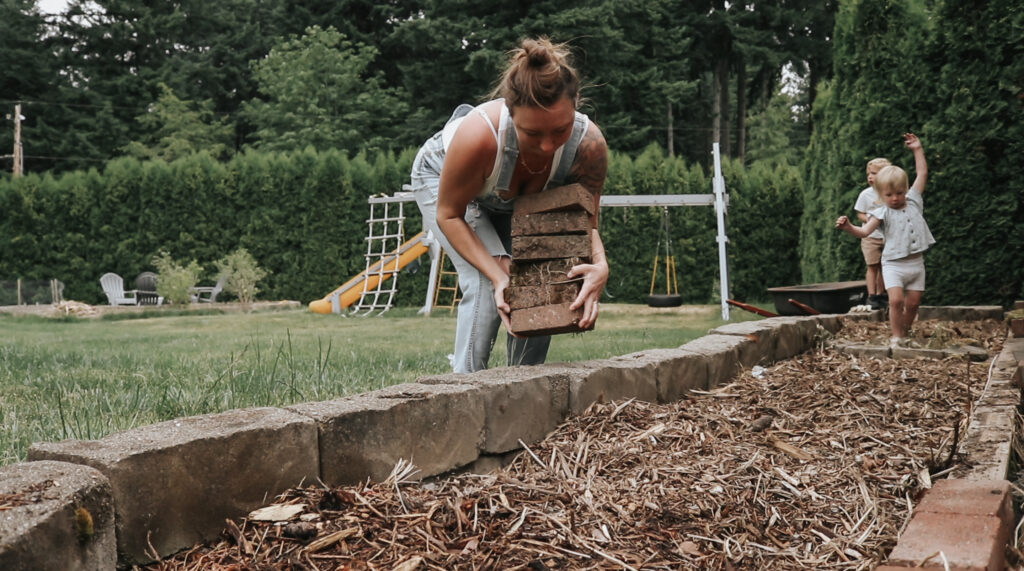 Christian homemaker wearing a white tank top and denim overalls carries a stack of bricks while working in a garden bed bordered by stone with children playing in the background.